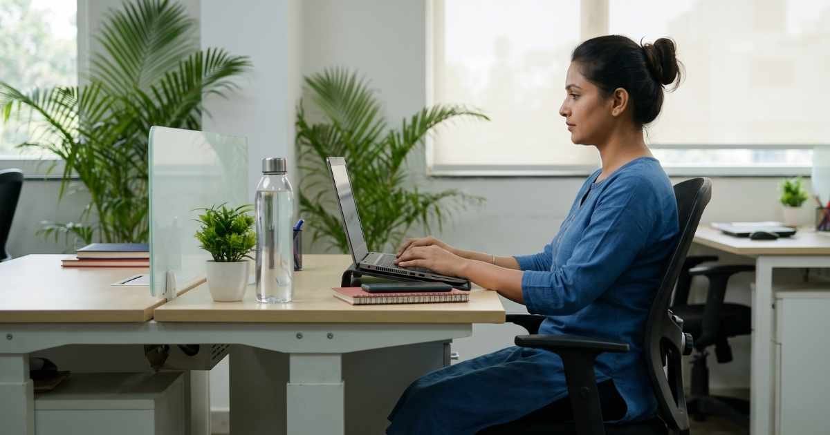 A woman sitting at an office desk working on a laptop with a straight back, supported neck, and proper ergonomic posture to prevent pain.